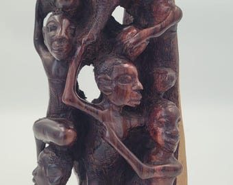 May include: A carved wooden sculpture showing several figures in a dark brown wood. The figures are intertwined, with visible facial and bodily details. The sculpture has a natural wood base and a smooth finish.