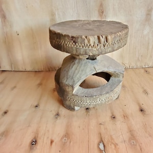 Puede incluir: Un taburete de madera con una parte superior redonda y una base tallada. El taburete está hecho de madera marrón oscura y tiene un aspecto rústico.