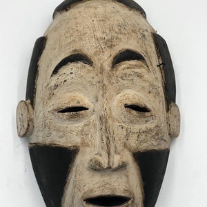 May include: A wooden mask with a carved human face. The mask is painted in shades of brown and white, with black accents around the eyes and mouth. The mask has a closed-eye expression and a slightly open mouth.