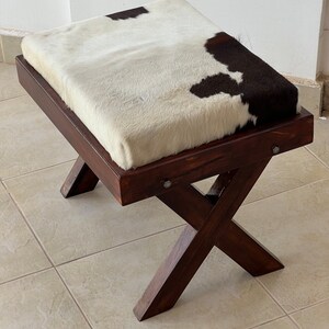 Könnte beinhalten: Ein Hocker aus Holz mit einer Sitzfläche aus weißem und schwarzem Kuhfell. Der Hocker hat ein X-förmiges Gestell.