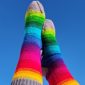 May include: A pair of colorful, knee-high socks against a clear blue sky. The socks display a vibrant rainbow pattern, transitioning from red to yellow, green, and blue. The cuffs and toes are a solid gray color, providing a contrasting detail.