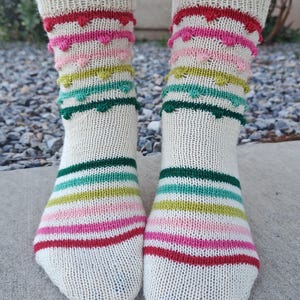 Puede incluir: Un par de calcetines de punto color crema con rayas horizontales rojas, rosas, verdes y turquesas. La parte superior de los calcetines presenta un diseño festoneado en colores coordinados. Los calcetines se muestran sobre una superficie de hormigón.