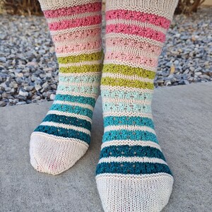 Puede incluir: Un par de calcetines tejidos a mano con puño y puntera beige. Los calcetines presentan rayas horizontales en rosa, verde y turquesa, con una sección estampada entre las rayas rosa y verde. Los calcetines están hechos de hilo suave.