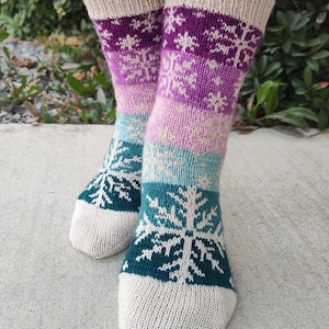 Könnte beinhalten: Ein Paar gestrickte Socken mit einem Schneeflockenmuster in den Farben Lila, Türkis und Weiß. Die Socken sind aus cremefarbenem Garn gefertigt.