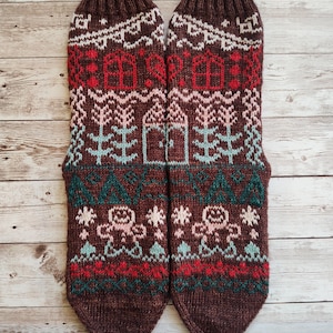 Op de afbeelding: Een paar bruine gebreide sokken met een kleurrijk winterthema. Het patroon bevat huizen, bomen, sneeuwvlokken en peperkoekmannetjes.