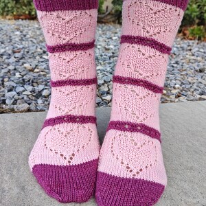 May include: A pair of pink and burgundy knitted socks with a heart pattern. The socks have a ribbed cuff and a contrasting color heel and toe.