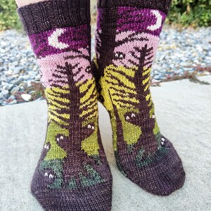 May include: A pair of knitted socks with a forest scene and a crescent moon. The socks are brown, purple, pink, yellow, and green.