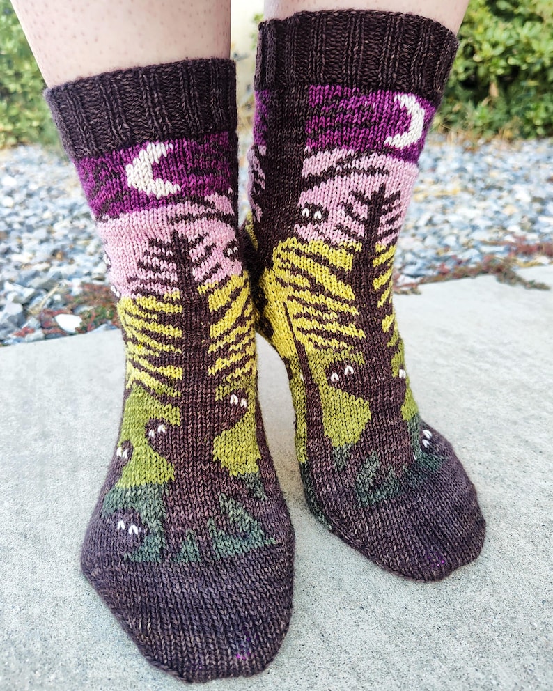 May include: A pair of knitted socks with a forest scene and a crescent moon. The socks are brown, purple, pink, yellow, and green.