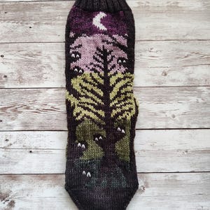 May include: A pair of knitted socks with a forest scene design. The socks are brown, green, purple, and white. The design features a tree, a crescent moon, and several eyes.