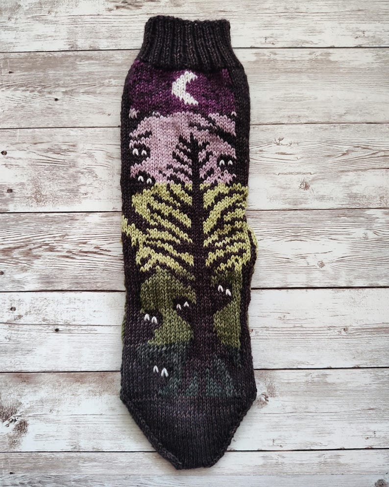 May include: A pair of knitted socks with a forest scene design. The socks are brown, green, purple, and white. The design features a tree, a crescent moon, and several eyes.