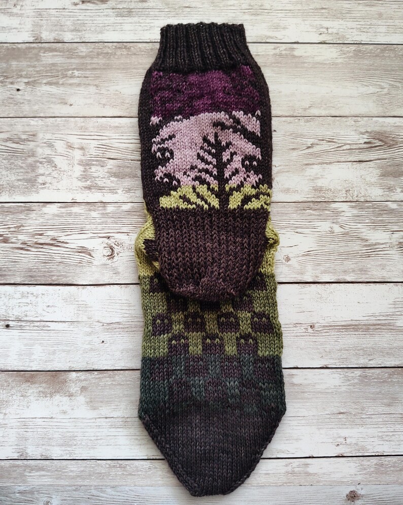 May include: A pair of knitted socks with a brown, green, and purple color scheme. The socks feature a forest scene with trees and a sunset.