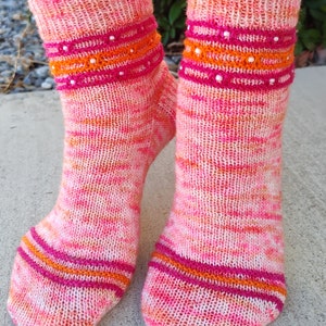 Puede incluir: Un par de calcetines de punto rosa, naranja y blanco con un patrón de rayas rosa y naranja y cuentas blancas alrededor de la parte superior de los calcetines.