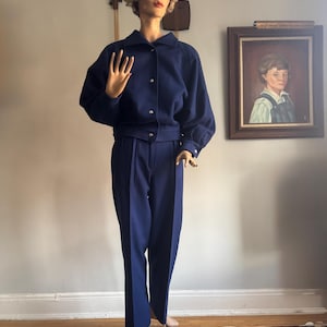 May include: A navy blue two-piece suit displayed on a mannequin. The jacket has a rounded collar and gold buttons. The pants are high-waisted with a pleated front. The mannequin is positioned in front of a neutral wall with a framed portrait.