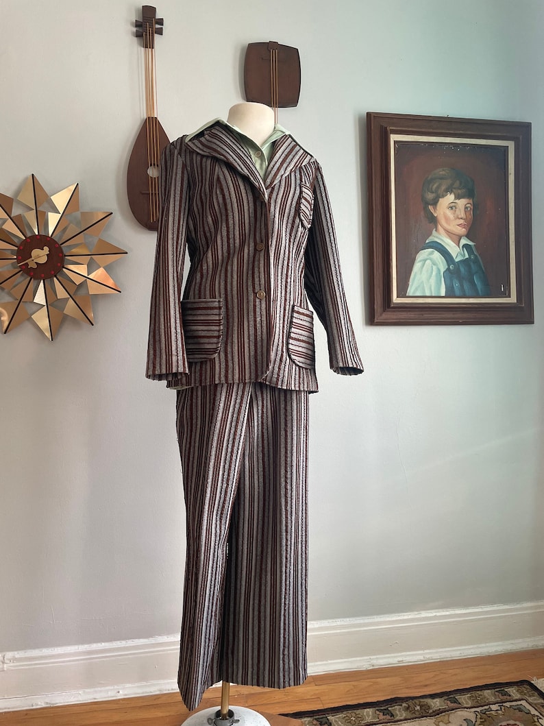 May include: A striped trouser suit in brown and grey with a long-sleeved jacket and wide-leg trousers. The jacket has a notched collar, a button fastening, and two flap pockets. The trousers have a straight leg and a zip fastening.