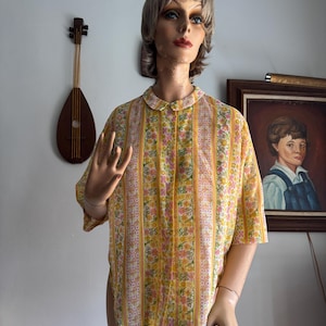 May include: A vintage short-sleeved shirt on a mannequin. The shirt features a yellow background with vertical stripes and floral patterns in pink, orange, and white. It has a rounded collar and a loose fit, typical of retro styles.