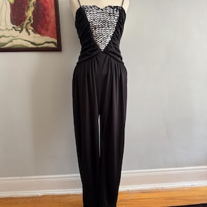 May include: A black jumpsuit with a silver sequined heart-shaped design on the bodice. The jumpsuit has thin shoulder straps and a wide-leg silhouette.