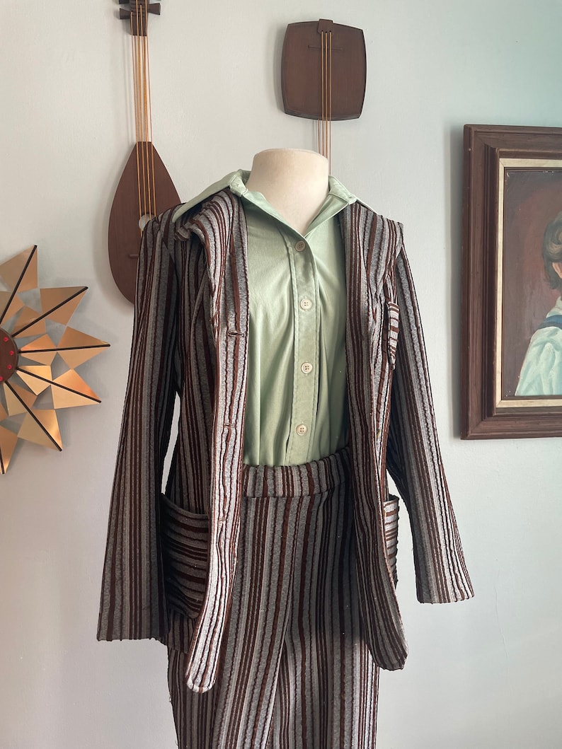 May include: A striped brown and grey trouser suit with a matching jacket and trousers. The jacket has a collar and pockets. The trousers have a high waist and a straight leg. The suit is on a mannequin and is displayed against a white wall.