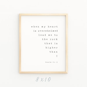 Pode incluir: Uma impressão emoldurada com fundo branco e texto preto. O texto diz "when my heart is overwhelmed lead me to the rock that is higher than I Psalm 61:2". A moldura é de madeira marrom claro e a impressão mede 20,32 x 25,4 cm.