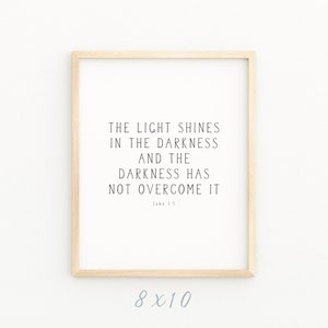 May include: A framed print with a white background and black text that reads "The light shines in the darkness and the darkness has not overcome it. John 1:5". The frame is light brown wood and the print is 8x10 inches.
