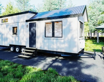 May include: A white tiny house on wheels with a black roof and a black door. The house has two windows on the side and a small window on the front. The house is parked on a gravel driveway with green grass and trees in the background.
