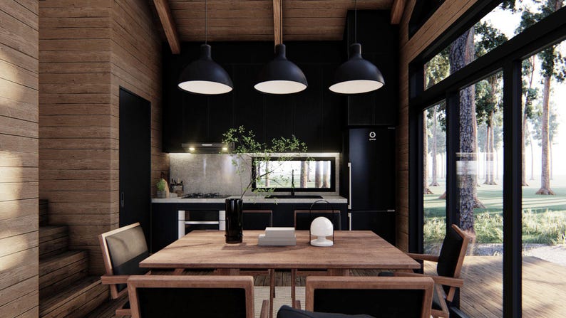 May include: Interior shot of a modern kitchen and dining area. The space features a wooden table and chairs, black pendant lights, and a black kitchen with a refrigerator. Large windows offer a view of trees and natural light.