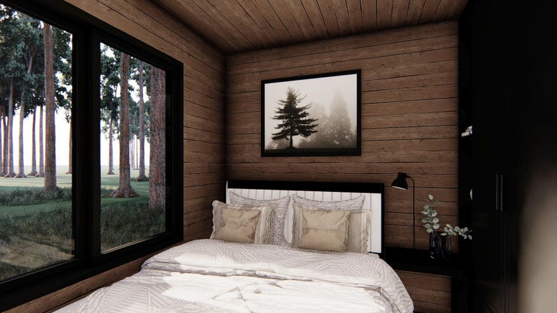 May include: A cozy bedroom with wood-paneled walls and ceiling. A large window offers a view of a forest. The bed has a white comforter and pillows. A framed picture of a tree hangs above the bed. A black nightstand and lamp are visible.