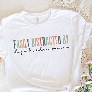 May include: White t-shirt with the text "EASILY DISTRACTED BY dogs & video games" in colorful letters.