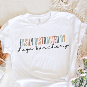 May include: White t-shirt featuring the phrase "EASILY DISTRACTED BY dogs & archery" in a colourful, whimsical font. The shirt is presented on a soft, neutral-toned surface, with a woven basket and flowers. The text is in a mix of colours.