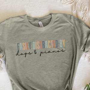 May include: A heather grey t-shirt with the text "EASILY DISTRACTED BY dogs & pianos" in colorful letters.