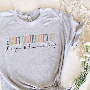 May include: A heather grey t-shirt with the text "EASILY DISTRACTED BY dogs & dancing" in colorful letters.
