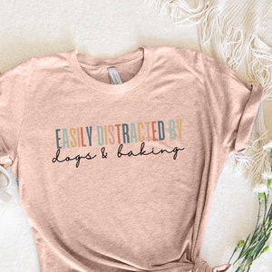 May include: A heather peach coloured t-shirt with the text "Easily Distracted by dogs & baking" in colourful lettering.