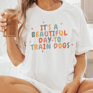 Pode incluir: Camiseta branca com o texto "It's a beautiful day to train dogs" em letras coloridas. A camiseta está sendo usada por uma pessoa sentada em uma bancada branca.