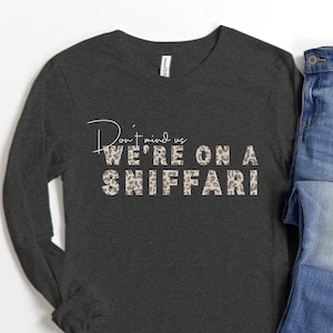 May include: A dark gray long-sleeve shirt with the text "Don't mind us WE'RE ON A SNIFFARI" in white with a leopard print pattern. The shirt is shown on a white background with a pair of blue jeans.