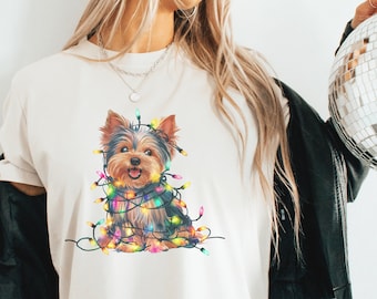 Może przedstawiać: Kremowy t-shirt z Yorkshire Terrierem owiniętym w kolorowe lampki choinkowe. Pies ma brązowo-czarną sierść i uśmiecha się. Koszulka ma swobodny styl.