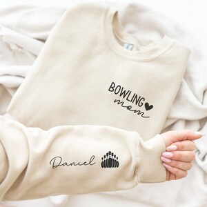 May include: Beige sweatshirt with the text "Bowling mom" and a heart on the front. The left sleeve has the text "Daniel" and a graphic of bowling pins.