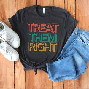 May include: A black t-shirt with the text "TREAT THEM RIGHT" in colorful block letters. The shirt is laid out on a wooden surface with a pair of white sneakers and blue jeans.