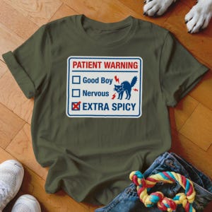 May include: Olive green t-shirt with a graphic that reads "PATIENT WARNING" above a checklist. The checklist includes "Good Boy," "Nervous," and "EXTRA SPICY" with the last option checked. A blue cat with lightning bolts is also featured.