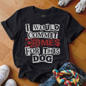 May include: Black t-shirt with the text "I WOULD COMMIT CRIMES FOR THIS DOG" in a distressed font. The word "CRIMES" is in red. The shirt is paired with blue jeans, a dog toy, and white sneakers.
