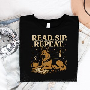 Könnte beinhalten: Schwarzes T-Shirt mit dem Text "READ. SIP. REPEAT." und einer Grafik eines Hundes, einer Kaffeetasse, eines Buches und einer Kerze. Der Hund ist braun und hellbraun, und die Tasse ist hellbraun. Das T-Shirt liegt auf einer Jeans.