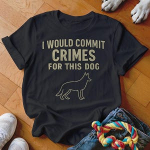 May include: Black t-shirt with the text "I WOULD COMMIT CRIMES FOR THIS DOG" in a light tan color. Below the text is a line drawing of a dog. The shirt is folded, with a pair of white sneakers, blue jeans, and a dog toy in the image.