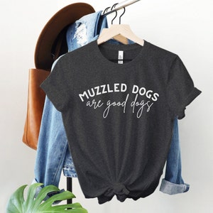 May include: A dark gray t-shirt with the text "MUZZLED DOGS are good dogs" in white lettering. The shirt is hanging on a hanger with a denim jacket and a brown leather bag in the background.