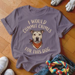 May include: A lilac-colored t-shirt with the text "I WOULD COMMIT CRIMES FOR THIS DOG" and a graphic of a dog wearing a red bandana. The shirt is folded, with a pair of white sneakers, blue jeans, and a dog toy in the image.