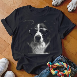 Könnte beinhalten: Schwarzes T-Shirt mit einem schwarz-weißen Bild eines Border Collies mit Pilotenbrille. Das Gesicht des Hundes steht im Mittelpunkt, mit sichtbaren Ohren und Brust. Das T-Shirt liegt auf einer Holzoberfläche mit anderen Gegenständen.