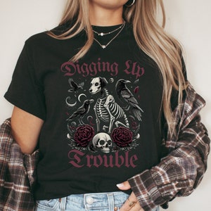 May include: Black t-shirt with a graphic design of a skeleton dog with crows and flowers. The text "Digging Up Trouble" is printed in a gothic font.