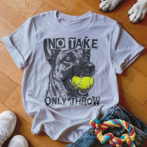 May include: White t-shirt with a graphic of a dog holding a yellow tennis ball in its mouth. The text "NO TAKE ONLY THROW" is printed above and below the dog illustration. The shirt is displayed with jeans, sneakers, and a dog toy.