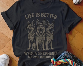 May include: Black t-shirt featuring a graphic of three German Shepherds. The text "LIFE IS BETTER WITH A SHEPHERD OR TWO, OR THREE" is printed above and below the dogs in a distressed font. The shirt is folded on a wooden surface.
