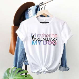 May include: White t-shirt with the text "I'd rather be training my dog" in colorful letters.