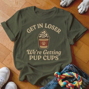 Puede incluir: Camiseta verde oliva con el texto "Get in Loser, We're Getting Pup Cups" y un gráfico de una taza de café con una huella de pata. La camiseta está doblada, con unos vaqueros, un juguete para perros y zapatillas blancas en la imagen.