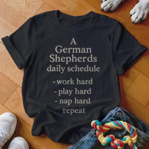 May include: Black t-shirt with the text "A German Shepherds daily schedule - work hard - play hard - nap hard - repeat" in a light grey font. The shirt is folded on a wooden surface, with a dog toy and jeans visible.