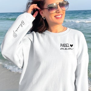 May include: White sweatshirt with the text "PADEL MOM" and a black heart on the front. The left sleeve has a black and white illustration of a paddle and the text "Padel" written in cursive.